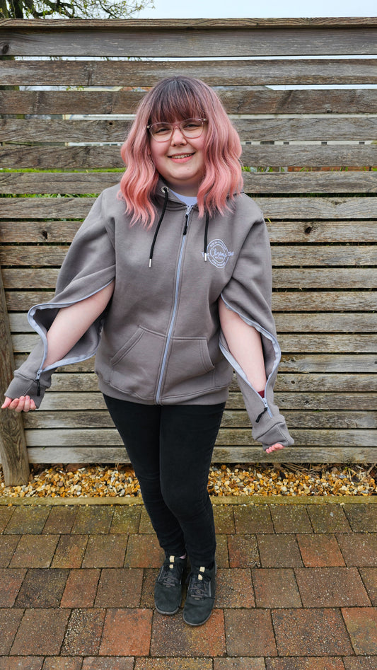 A young white woman with ombre brown to pink hair, stood against a wooden fence, wearing an IV access hoodie. She has zips in both arms which are undone, exposing her skin for IV access. She is smiling.