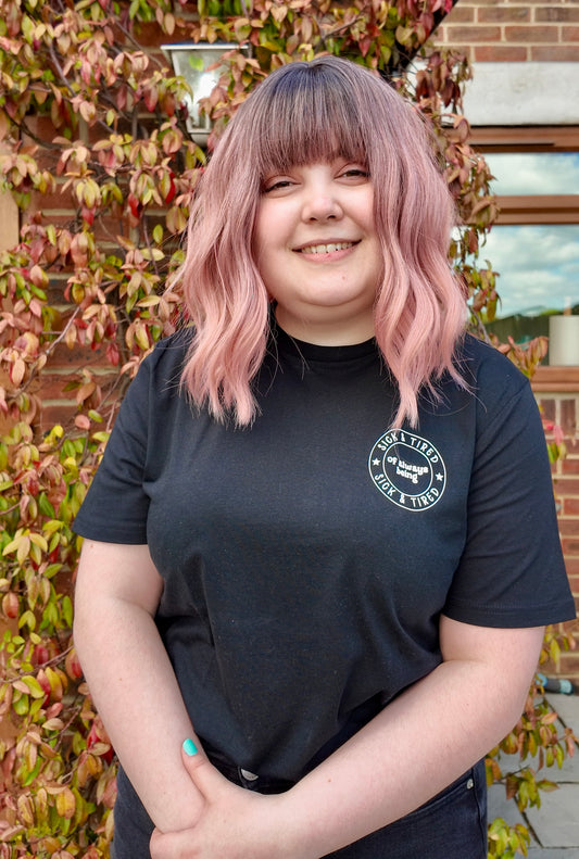 A woman with brown to pink ombre hair, wearing a black t-shirt that says 'sick and tired of always being sick and tired'. She is smiling at the camera 