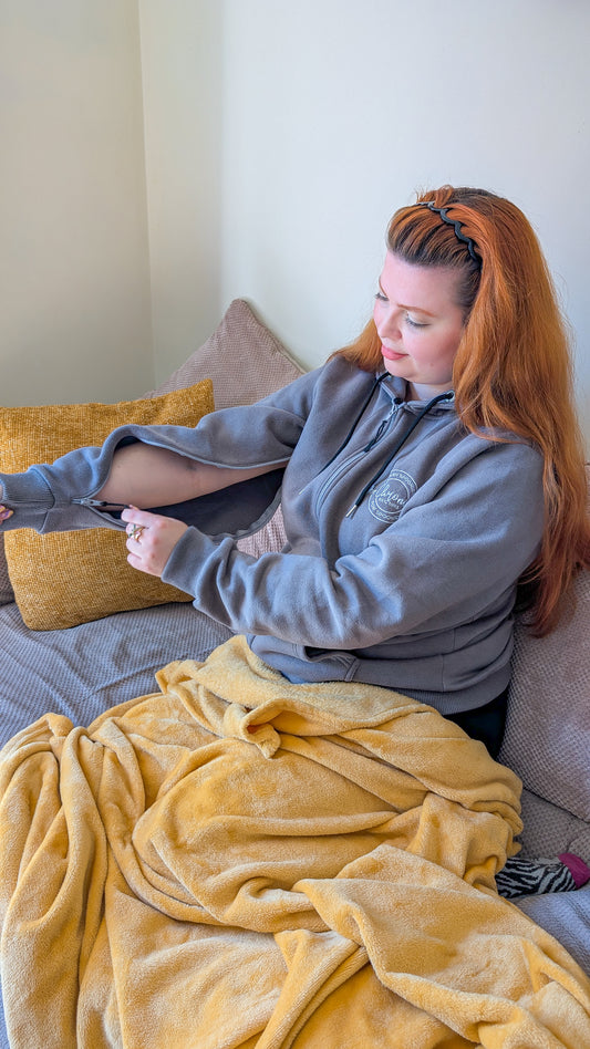 A woman with red hair, wearing a grey IV access hoodie. She is sitting on a sofa with an mustard yellow blanket on her lap.