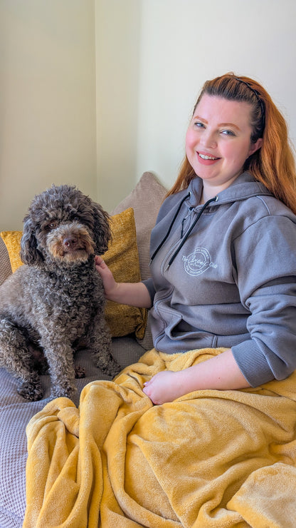 A woman with red hair, wearing a grey IV access hoodie. She is sitting on a sofa with an mustard yellow blanket on her lap. Next to her is a curly, brown haired dog.
