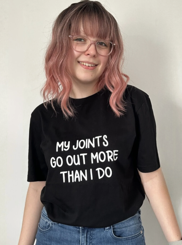 Woman with pink hair wearing a black top that says 'My joints go out more than I do'