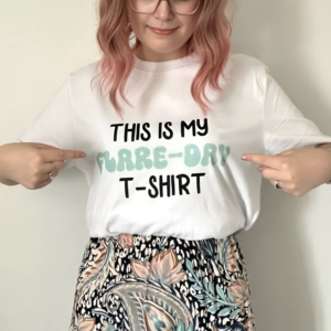 Woman with pink hair, wearing a white top which she is pointing at, which says 'this is my flare-day t-shirt'