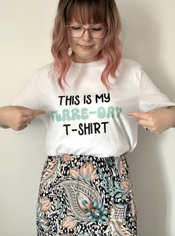 Woman with pink hair, wearing a white top which she is pointing at, which says 'this is my flare-day t-shirt'